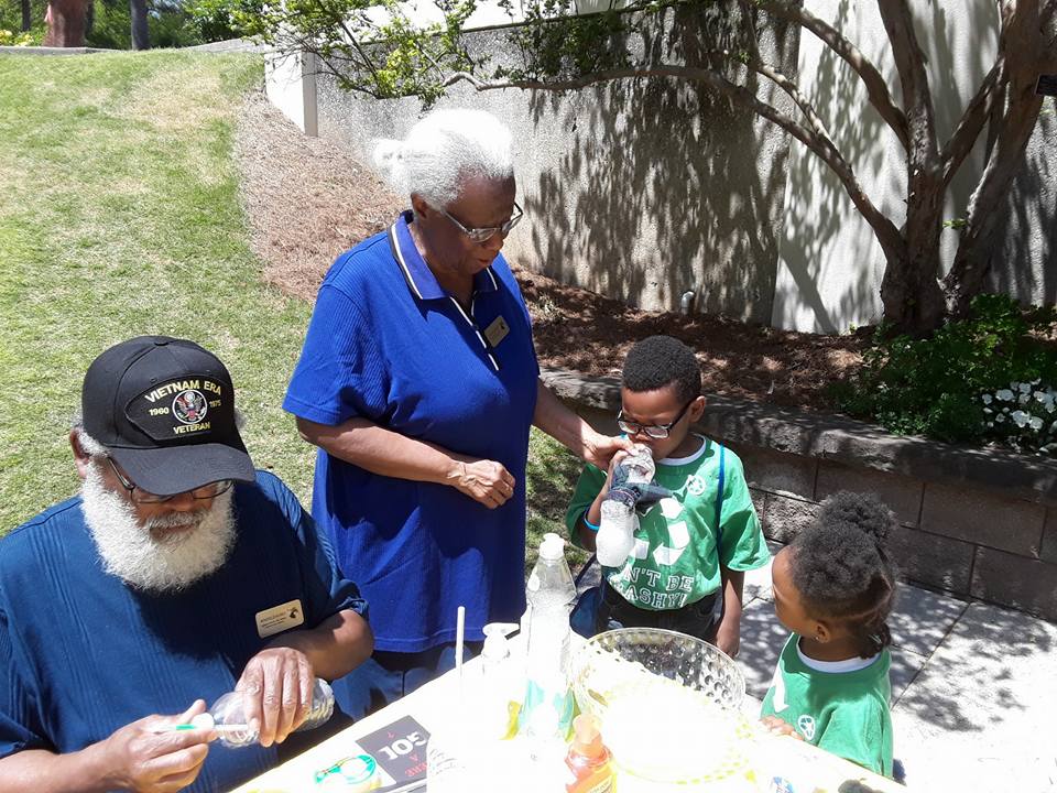 Photo shows the Burks wearing blue shirts leading an environmental science lesson with two small children wearing green shirts on Earth Day at Birmingham Botanical Gardens in Alabama