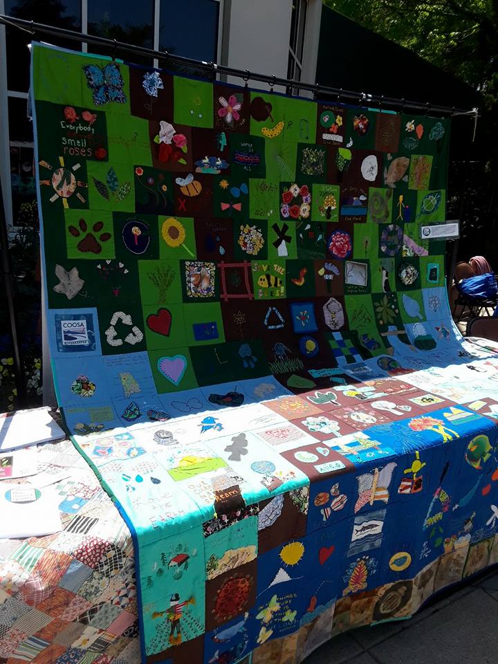 Photo shows a quilt-covered table. A large community quilt created from felt squares by Earth Day attendees at Birmingham Botanical Gardens hangs above it.