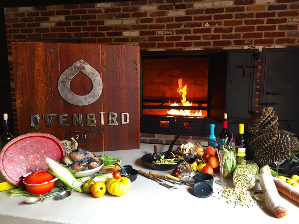 Photo shows a brick hearth with fire, a table full of fresh produce and an wooden plaque bearing the logo for OvenBird, a restaurant based in the Pepper Place district in Birmingham, Alabama