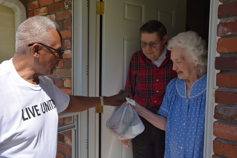 A man greets an elderly couple at their door with a warm meal
