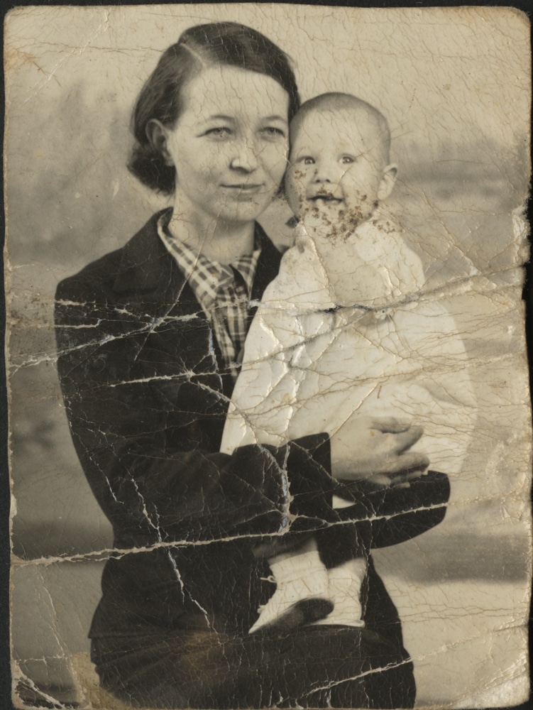An old photo with crack and discoloration. It pictures a woman holding a child.
