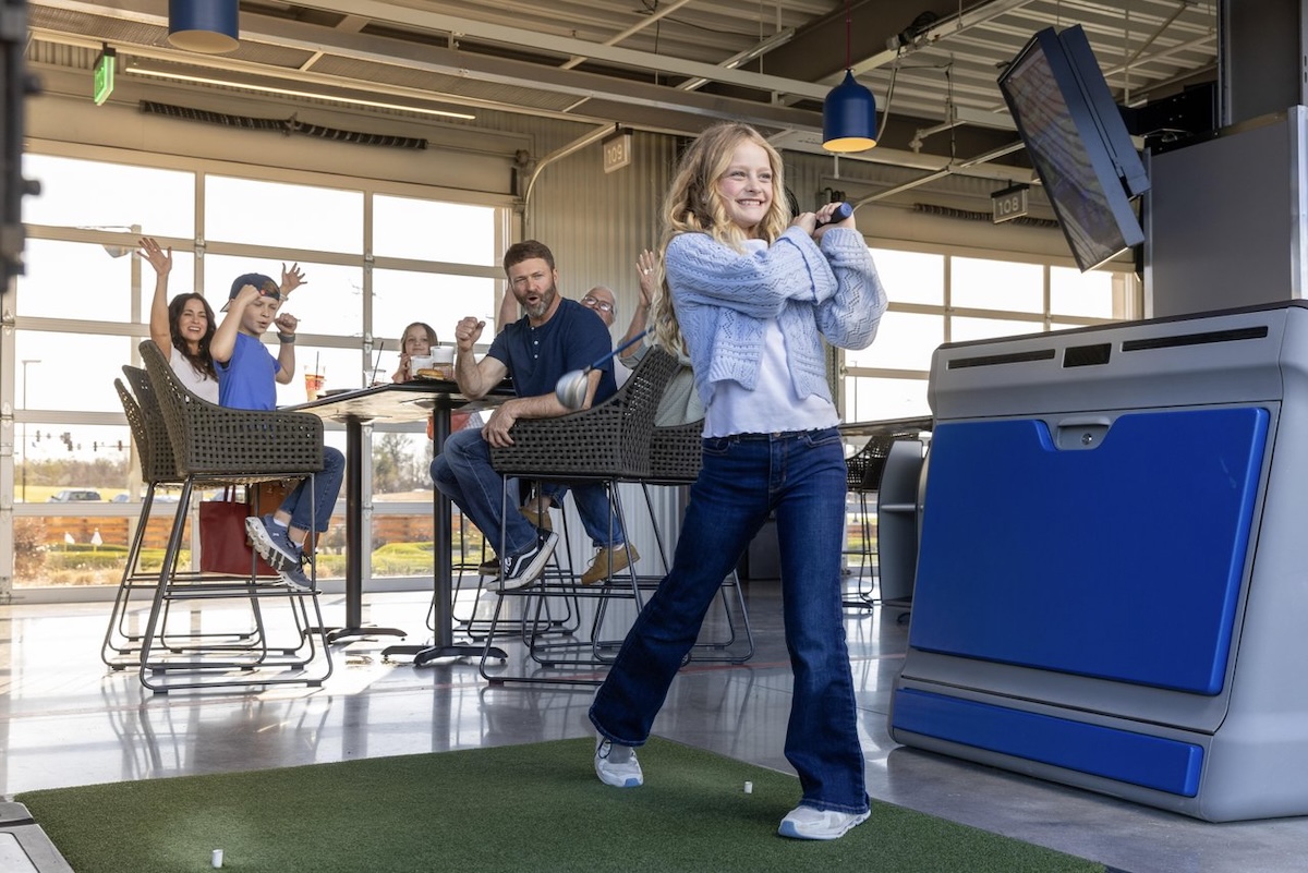 A young girl takes a swing at a golf ball at Top Golf in Ridgeland, Mississippi, while her family cheers her on in the background.