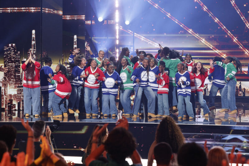 a large group of young people of many ages in blue, red and green jerseys celebrates with one another on the americas got talent stage